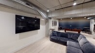 This single-floor amenity space combines industrial design elements with modern comforts to create a dynamic and inviting environment for gathering and recreation. Exposed concrete ceilings with visible ductwork and structural columns lend an authentic industrial character throughout, while light hardwood flooring and crisp white walls keep the atmosphere clean and contemporary.

The main lounge area serves as the centerpiece of the space, featuring a generous L-shaped sectional sofa in deep navy upholstery positioned in front of a large wall-mounted flat-screen television. Nearby, high-top bar tables and stools provide additional casual seating options. Framed artwork accents the wall, adding a polished touch to the industrial backdrop.

Adjacent to the lounge, a dedicated billiards zone houses a full-size pool table with a rich wood frame and blue felt surface, complemented by a mounted cue rack and high-top seating—perfect for friendly competition.

The well-appointed kitchenette features dark cabinetry with white countertops, a textured tile backsplash, stainless steel appliances including a full-size refrigerator, microwave, and sink, providing all the essentials for hosting and entertaining.

A separate enclosed room, visible through a glass panel, offers a more intimate setting with its own seating arrangement, ideal for private gatherings or quieter conversations. Together, these areas create a versatile, amenity-rich space designed for community engagement.