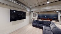 This single-floor amenity space combines industrial design elements with modern comforts to create a dynamic and inviting environment for gathering and recreation. Exposed concrete ceilings with visible ductwork and structural columns lend an authentic industrial character throughout, while light hardwood flooring and crisp white walls keep the atmosphere clean and contemporary.

The main lounge area serves as the centerpiece of the space, featuring a generous L-shaped sectional sofa in deep navy upholstery positioned in front of a large wall-mounted flat-screen television. Nearby, high-top bar tables and stools provide additional casual seating options. Framed artwork accents the wall, adding a polished touch to the industrial backdrop.

Adjacent to the lounge, a dedicated billiards zone houses a full-size pool table with a rich wood frame and blue felt surface, complemented by a mounted cue rack and high-top seating—perfect for friendly competition.

The well-appointed kitchenette features dark cabinetry with white countertops, a textured tile backsplash, stainless steel appliances including a full-size refrigerator, microwave, and sink, providing all the essentials for hosting and entertaining.

A separate enclosed room, visible through a glass panel, offers a more intimate setting with its own seating arrangement, ideal for private gatherings or quieter conversations. Together, these areas create a versatile, amenity-rich space designed for community engagement.