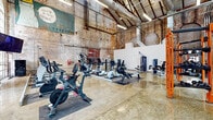 Located in Memphis, TN, this fitness center occupies a generously sized single-floor space that masterfully blends historic industrial architecture with modern gym amenities. The building's original exposed brick walls, adorned with eclectic murals and graffiti-style artwork, lend the space a distinctive urban character that sets it apart from conventional fitness facilities. Soaring vaulted ceilings with visible ductwork and industrial lighting fixtures amplify the sense of openness, while large arched windows along the upper walls flood the interior with abundant natural light.

The polished concrete floors provide a durable, reflective surface that enhances brightness throughout the space. The fitness floor is thoughtfully organized to accommodate a wide range of workout styles. Cardio equipment — including treadmills, ellipticals, stationary bikes, and rowing machines — is arranged along one section, each station accompanied by mounted display screens. A dedicated strength training zone features an orange-accented multi-functional power rack with an extensive dumbbell selection. Selectorized weight machines are positioned nearby for targeted muscle training. A standout feature is the colorful climbing wall mounted near the large glass entry doors, offering a dynamic functional fitness option. The open layout ensures ample room to move freely between stations, creating an energizing and well-equipped workout environment.