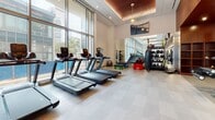 This contemporary fitness center is designed to offer a premium workout experience within a thoughtfully appointed space. The facility is defined by soaring floor-to-ceiling glass windows that line the perimeter, flooding the interior with abundant natural light and providing open views of the surrounding urban landscape. The high ceilings feature elegant recessed wood panel accents with integrated linear and pendant lighting, creating a warm yet modern ambiance that complements the clean, light-toned flooring throughout.

The cardio zone is generously outfitted with multiple treadmills, elliptical machines, and a StairMaster, all positioned to take full advantage of the expansive window views. Adjacent to this area, the strength training section provides a well-rounded selection of equipment, including cable machines, resistance machines, adjustable benches, and a dedicated free weights area. A heavy bag and functional training rack round out the facility's functional fitness offerings.

Full-length mirrors are strategically placed along the walls, enhancing the sense of space and supporting proper form during workouts. The open, single-floor layout ensures a seamless flow between training zones, making the facility practical and user-friendly. Overall, the fitness center strikes a balance between high-end design and comprehensive functionality.
