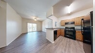 This single-floor residence offers four bedrooms, two full bathrooms, and a thoughtfully designed layout that maximizes functionality throughout. The open-concept living and kitchen area serves as the heart of the home, featuring wood-style laminate flooring, vaulted ceilings, and a ceiling fan. A sliding glass door floods the living area with natural light and opens to a private balcony with outdoor views. The kitchen is equipped with warm-toned wooden cabinetry, dark countertops, black appliances including a range, dishwasher, and refrigerator, and a peninsula that connects seamlessly to the living space. A central hallway connects the bedrooms and bathrooms, maintaining a clean and organized flow. The bedrooms are carpeted and finished with neutral tones, offering versatile, comfortable spaces. Three closets provide ample storage throughout. Both bathrooms feature wood-style flooring, warm wooden vanity cabinetry, white countertops, and full tub-shower combinations. The overall aesthetic is clean and neutral, with a consistent warm palette of beige walls and light wood tones creating a welcoming and cohesive ambiance throughout the home.