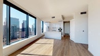 This contemporary high-rise residence is defined by clean lines, a bright neutral palette, and an abundance of natural light streaming through large floor-to-ceiling windows that frame expansive city skyline views. Light-toned hardwood flooring runs consistently throughout, unifying the open-concept layout.

The kitchen is a standout feature, showcasing sleek flat-front white cabinetry extending to the ceiling, a gas cooktop, stainless steel double wall ovens, a refrigerator, and a generous central island with a sink and ample counter space — all finished with crisp white surfaces for a polished, modern aesthetic.

The living area flows seamlessly from the kitchen, bathed in natural light and offering stunning urban vistas. A distinctive curved structural column adds architectural character to the main living space.

The bedrooms are bright and airy, each benefiting from large windows with elevated city views and warm hardwood flooring. A clean, well-lit hallway connects the private spaces.

The bathrooms are finished to a high standard: one features a soaking tub with an overhead rain shower set against all-white tile surrounds, while the primary bathroom offers a tub with a frameless glass partition and large-format tile work. The overall ambiance is sophisticated, minimal, and urban.