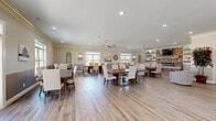 Located at 820 South Gateway Plaza Boulevard in Clarksville, Tennessee, this single-story community clubhouse offers a generous, open-concept layout designed for gathering and socializing. The main area is anchored by multiple large farmhouse-style dining tables with upholstered, nail-head-trim chairs, providing ample communal seating throughout the space. Light hardwood-style flooring runs continuously underfoot, complementing the warm, neutral color palette of the walls and décor.

A standout feature is the full-service kitchen, appointed with gray shaker-style cabinetry, white quartz countertops, subway tile backsplash, stainless steel appliances, a large center island, and open floating shelves. A picture window over the sink fills the kitchen with natural light and views of the surrounding community.

The lounge area offers a comfortable sectional sofa, accent chairs, and a striking floor-to-ceiling brick fireplace with a wall-mounted television and rustic floating shelves. Crown molding and two-tone wainscoting walls add architectural refinement throughout.

Large windows on multiple sides of the building invite abundant natural light and outdoor views, while ceiling fans provide additional comfort. Recessed lighting and pendant fixtures ensure the space is well-illuminated at all hours, creating a welcoming and functional environment for residents.