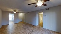 This single-floor space is designed with a straightforward, functional layout that maximizes usability. The main living area is finished with light-toned hardwood-style flooring that extends throughout, creating a clean, cohesive look across the space. A textured ceiling adds subtle visual interest overhead, while two ceiling fans with integrated light fixtures provide both air circulation and ambient lighting to the open area.

The neutral gray walls offer a versatile backdrop, complementing the light flooring and white trim. Multiple doorways are visible from the main living area, indicating a well-connected floor plan. One doorway opens to a hallway with additional closed doors, likely leading to bedrooms or storage areas, while another doorway provides a direct view into a bathroom, which features light-colored walls and accessible fixtures, including a grab bar.

A thermostat is mounted on the wall, and light switches are conveniently positioned throughout. The overall ambiance is bright, clean, and move-in ready, with a practical design that suits a variety of living arrangements. The open feel of the main area and the continuity of the flooring contribute to a sense of spaciousness throughout the unit.