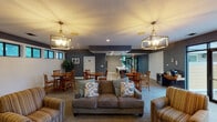 This single-floor community clubhouse offers a welcoming and well-appointed gathering space designed for both relaxation and social interaction. The main lounge area is generously proportioned, featuring plush seating including a sofa and striped armchairs arranged around a central conversation area. Two elegant gold-toned geometric pendant chandeliers illuminate the space, complementing the warm, carpeted flooring beneath. The walls are finished in deep navy and gray tones, accented by crisp white shiplap paneling that lines the lower portion of one wall. A continuous row of clerestory windows above the shiplap admits abundant natural light and offers views of the surrounding greenery, enhancing the open, airy feel of the room. Multiple round wooden dining tables with ladder-back chairs are arranged throughout the space, providing ample seating for group dining or casual gatherings. The kitchen area is equipped with white cabinetry, a stainless steel refrigerator, a sink, and a striking large island topped with marble-style countertops, lit by industrial-style pendant lights. Large glass sliding doors at one end of the space open directly to an outdoor patio area, seamlessly connecting the interior to the exterior and flooding the room with additional natural light. A beverage station and wall-mounted television round out the functional amenities of this versatile community space.