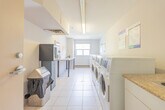 Located at 75 Huron Heights Dr in Newmarket, Ontario, this shared laundry facility is a compact, well-organized space designed to serve building residents efficiently. The room features a row of commercial-grade front-loading washers and top-loading dryers lined along one wall, offering ample laundry capacity. A granite-topped folding table runs along the opposite wall, providing a convenient surface for sorting and folding laundry. The floor is finished with light-coloured ceramic tiles that are easy to maintain and contribute to the room's clean, functional aesthetic. A window positioned at the far end of the room allows natural light to brighten the space while offering a view of the surrounding neighbourhood. Overhead fluorescent lighting ensures the room remains well-lit at all times. The textured ceiling, ventilation grilles, and a wall-mounted payment kiosk reflect the practical, utility-focused design of the space. Informational signage and a bulletin board are posted on the walls for resident reference. A fire extinguisher is visibly mounted near the entrance, ensuring safety compliance. The space is accessed through a solid door and maintains a tidy, straightforward layout suited to its shared-use purpose.