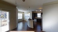 This single-story space is designed with an open-concept layout that seamlessly connects the kitchen and living areas. The kitchen is a functional centerpiece, featuring dark cabinetry that contrasts sharply with a white island topped with granite-style countertops. Track lighting illuminates the kitchen workspace, while pendant lights hang above the island, adding a layered lighting scheme. Stainless steel appliances, including a refrigerator, complement the dark cabinetry, and a window above the sink brings in natural light. The living area extends from the kitchen, benefiting from multiple large windows fitted with horizontal blinds that allow ample daylight to fill the space. Rich, dark hardwood flooring runs consistently throughout, grounding the interior with warmth and visual continuity. Crown molding along the ceilings adds a refined architectural detail to the common areas. A long hallway connects additional areas of the unit, lined with the same dark hardwood floors and accented by crown molding. Natural light filters through a window at the hallway's end, creating a bright and welcoming passage through the space. The overall ambiance is clean, modern, and well-lit.