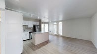This single-floor space showcases an open-concept layout that seamlessly connects the kitchen and living areas, creating a spacious and airy atmosphere. The kitchen is outfitted with crisp white cabinetry, granite countertops, and stainless steel appliances, including a refrigerator and range. A central island with a matching granite surface provides additional workspace and serves as a natural divider between the kitchen and the open living area. Track lighting runs along the ceiling, offering versatile and even illumination throughout the space. The living area is defined by a striking white-painted exposed brick accent wall, adding a layer of texture and character to the otherwise clean, modern aesthetic. Two arched windows allow natural light to filter in, enhancing the bright and welcoming ambiance. Light-toned hardwood-style flooring flows continuously throughout the space, further reinforcing the cohesive, open feel. Large windows at the rear of the kitchen bring in additional natural light, making the entire space feel open and connected to the outdoors. The overall design balances modern finishes with industrial-inspired details.
