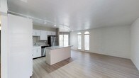 This single-floor space showcases an open-concept layout that seamlessly connects the kitchen and living areas, creating a spacious and airy atmosphere. The kitchen is outfitted with crisp white cabinetry, granite countertops, and stainless steel appliances, including a refrigerator and range. A central island with a matching granite surface provides additional workspace and serves as a natural divider between the kitchen and the open living area. Track lighting runs along the ceiling, offering versatile and even illumination throughout the space. The living area is defined by a striking white-painted exposed brick accent wall, adding a layer of texture and character to the otherwise clean, modern aesthetic. Two arched windows allow natural light to filter in, enhancing the bright and welcoming ambiance. Light-toned hardwood-style flooring flows continuously throughout the space, further reinforcing the cohesive, open feel. Large windows at the rear of the kitchen bring in additional natural light, making the entire space feel open and connected to the outdoors. The overall design balances modern finishes with industrial-inspired details.