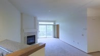 This single-floor residence offers a clean, open layout designed for comfortable everyday living. The main living area is anchored by a white-surround fireplace, adding a classic focal point to the room. Soft, wall-to-wall carpeting runs throughout the space, contributing to a warm and inviting atmosphere. Natural light pours in generously through a large sliding glass door paired with a transom window above, offering direct access to a private balcony that overlooks lush, verdant greenery — a serene backdrop that brings the outdoors in. The neutral color palette of white and soft beige walls provides a versatile canvas adaptable to a variety of interior styles. A partial partition with a wooden countertop edge defines the transition between the living area and the kitchen, suggesting an open-concept flow between the two spaces. The kitchen counter features a granite or stone surface, hinting at quality finishes throughout. Recessed lighting in the ceiling keeps the space feeling modern and uncluttered. The overall ambiance is calm, airy, and move-in ready, with a layout that balances openness with defined functional zones.