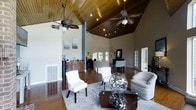 This single-floor space makes a striking first impression with its dramatic vaulted ceilings finished in rich, warm-toned wood paneling that draws the eye upward and creates an expansive, airy atmosphere. Decorative tropical-style ceiling fans are mounted throughout, adding both visual interest and practical comfort. The open-concept layout connects multiple functional areas seamlessly, allowing natural light to flow freely across the gleaming hardwood floors. A comfortable lounge area anchors the main space, featuring neutral-toned upholstered seating arranged around a glass-top coffee table accented with sculptural metallic orb décor. Abstract artwork adorns the tall cream-colored walls, adding a contemporary artistic touch. Toward the rear, a wet bar or kitchenette with wooden cabinetry and a countertop water dispenser provides convenient amenities. Beyond it, a secondary seating or workstation area is bathed in natural light from large windows that offer views to the outdoors. French doors with glass panes provide additional access points, enhancing the sense of openness. Structural columns punctuate the space without interrupting the flow, while recessed and track lighting ensures the interior is well-lit throughout. The overall ambiance is welcoming, polished, and versatile.