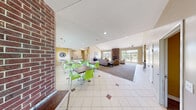 Located at 5825 Emerald Springs in Detroit, Michigan, this single-floor community space offers a welcoming and versatile layout designed for gathering and recreation. The main living area impresses with soaring vaulted ceilings, a striking floor-to-ceiling brick fireplace, and large glass doors that flood the room with natural light and provide views of the surrounding landscape. Comfortable seating, a wall-mounted television, and a built-in counter workspace make this area ideal for both relaxation and productivity. An adjacent dining nook features vibrant green seating and tile flooring, seamlessly transitioning between carpet and tile throughout the space. The second living area serves as a multipurpose room, equipped with a ceiling-mounted projector, a projection screen, plush tan leather seating, and a foosball table, making it well-suited for entertainment and community events. A community banner adds a personalized touch to the room. The kitchen is appointed with rich cherry wood cabinetry, dark granite countertops, a double sink, and tile flooring, providing ample functionality for catering and communal use. A connecting hallway ties the spaces together, completing this well-organized and inviting community facility.