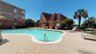 This outdoor amenity space centers around a generously sized freeform swimming pool with crystal-clear turquoise water, reaching a depth of 5 feet 6 inches. The pool is framed by an expansive brick-paver deck that provides ample room for lounging and socializing under open skies. Multiple chaise lounge chairs and shaded umbrella seating are positioned throughout the deck, offering comfortable options for relaxation in full sun or shade.

The standout architectural feature of this space is the adjacent clubhouse building, constructed of warm red brick with a steeply pitched, multi-gabled dark roof and distinctive arched windows that lend the structure a refined, traditional character. The pool area is fully enclosed by a black wrought iron fence with secured gated access points, ensuring both safety and privacy.

Surrounding multi-story residential buildings clad in brick and stucco rise alongside the pool area, providing a sheltered setting. Mature trees and tropical palm trees punctuate the landscape, adding greenery and a relaxed outdoor ambiance. A children's playground is also visible within the fenced grounds. The combination of the elegant clubhouse, well-maintained pool, and thoughtfully designed deck creates an inviting outdoor retreat ideal for leisure and community gathering.