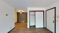 This single-floor residential space offers a straightforward, functional layout with consistent hardwood-style flooring that runs throughout, lending a warm and cohesive feel to the interior. The main living area is finished with neutral-toned walls and features a ceiling fan with a light fixture, providing both air circulation and ambient lighting. A baseboard heater along the far wall ensures efficient climate control throughout the colder months. The space includes a built-in closet fitted with classic white louvered sliding doors framed by dark wood trim, offering generous storage and a clean, traditional aesthetic. The open sightline between rooms creates a sense of connected space, making the floor plan feel open and accessible. White ceilings and light-colored walls help reflect available light, brightening the interior. The dark wood trim detailing around doorways and the closet adds a grounding contrast to the lighter palette of the walls and ceilings. Overall, the space presents a clean, move-in-ready environment with practical features suited for comfortable everyday living.