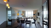 This single-floor residence showcases a thoughtfully designed open-plan layout that seamlessly integrates the living and dining areas. The space is anchored by dramatic floor-to-ceiling corner windows that flood the interior with natural light while framing sweeping panoramic views of the surrounding landscape, creating a bright and airy atmosphere throughout.

The dining area features a sleek, dark-toned table accompanied by elegant upholstered chairs with refined detailing, positioned to take advantage of the views. The living area is furnished with a neutral-toned sofa and accent chairs, complemented by carefully placed table lamps that add warmth to the space. A wall-mounted flat-screen television is integrated into the layout alongside a console table with decorative accessories.

Artwork adorns the walls, including a colorful abstract piece that adds a vibrant focal point. The dark hardwood flooring runs throughout, providing a rich contrast to the light-toned walls. The overall ambiance is sophisticated and contemporary, with a careful balance of natural light and curated furnishings that lend the space an elevated, polished character.