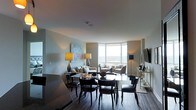 This single-floor residence showcases a thoughtfully designed open-plan layout that seamlessly integrates the living and dining areas. The space is anchored by dramatic floor-to-ceiling corner windows that flood the interior with natural light while framing sweeping panoramic views of the surrounding landscape, creating a bright and airy atmosphere throughout.

The dining area features a sleek, dark-toned table accompanied by elegant upholstered chairs with refined detailing, positioned to take advantage of the views. The living area is furnished with a neutral-toned sofa and accent chairs, complemented by carefully placed table lamps that add warmth to the space. A wall-mounted flat-screen television is integrated into the layout alongside a console table with decorative accessories.

Artwork adorns the walls, including a colorful abstract piece that adds a vibrant focal point. The dark hardwood flooring runs throughout, providing a rich contrast to the light-toned walls. The overall ambiance is sophisticated and contemporary, with a careful balance of natural light and curated furnishings that lend the space an elevated, polished character.