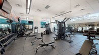 Located at 1750 Chestnut Pl in Denver, Colorado, this fitness center is a well-appointed, single-floor facility designed to support a comprehensive workout experience. The space is divided into distinct functional zones, each thoughtfully equipped and finished.

The main strength training area features rubber flooring and an extensive selection of cable machines, multi-functional cable systems, free weights, adjustable benches, and dumbbell racks. Full-length mirrors line the walls, enhancing the sense of openness throughout. A bold, colorful mural serves as a striking visual focal point, adding an energetic artistic element to the environment. A curved reception desk anchors the entrance to the strength area.

The cardio zone is positioned along a wall of floor-to-ceiling windows that flood the space with abundant natural light and offer views of the surrounding Denver skyline. A long row of treadmills, upright bikes, and cycling machines are arranged to take full advantage of these expansive urban views. The elevated clerestory windows above further amplify the brightness of the space.

A separate studio room provides a dedicated area for group fitness, stretching, or personal training, featuring hardwood-style flooring, stability balls, resistance bands, and framed artwork. A vibrant mural continues the artistic theme into this space as well.

Throughout, the facility maintains a clean, modern aesthetic with recessed lighting, a neutral white palette accented by teal, and a spacious layout that ensures comfortable circulation between equipment zones.