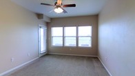 This single-story space presents a clean, functional layout centered around a well-lit main room. The room features wall-to-wall carpeting in a neutral tone, contributing to a warm and comfortable atmosphere. Smooth, beige-painted walls complement the understated aesthetic throughout.

Natural light is a defining characteristic of this space, with three evenly spaced windows along one wall providing ample brightness and views of the surrounding greenery. Each window is fitted with white horizontal blinds, offering flexible light control. A glass-paneled door on the adjacent wall further enhances the flow of natural light while providing convenient outdoor access.

A ceiling fan with an integrated light fixture is centrally mounted, ensuring both air circulation and overhead illumination. White baseboards provide a crisp contrast against the carpeted flooring and neutral walls, adding a clean, finished look to the room.

The overall ambiance is open and airy, making the space well-suited for a variety of furniture arrangements. The combination of natural light, soft carpeting, and neutral tones creates an inviting environment that is both practical and comfortable for everyday use.