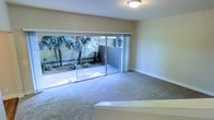 This single-story space offers a clean, functional layout highlighted by a generously sized living area finished with neutral-toned wall-to-wall carpeting. The room's most prominent feature is a large sliding glass door system that spans much of one wall, flooding the interior with natural light and providing direct access to a private outdoor patio. The patio is enclosed by a perimeter wall and framed by mature trees and established landscaping, creating a sense of seclusion and a natural backdrop visible from within the room. The mirrored sliding closet doors on the adjacent wall further amplify the sense of light and space, reflecting both the interior and the outdoor greenery. Walls are finished in a soft, warm neutral tone that complements the carpeting, contributing to a calm and understated ambiance. A flush-mount ceiling light fixture provides overhead illumination. The overall design is straightforward and practical, emphasizing the connection between the interior living space and the sheltered outdoor area, making it well-suited for those who value privacy alongside access to natural surroundings.