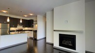 This single-floor space showcases a clean, modern design with an open-concept layout that seamlessly connects the living and kitchen areas. The living space is anchored by a striking floor-to-ceiling white surround fireplace with a minimalist metal shelf, serving as a bold focal point against the crisp, neutral walls. Dark hardwood flooring runs throughout, adding warmth and contrast to the light-toned walls and creating a cohesive aesthetic.

The kitchen features dark espresso cabinetry paired with white lower cabinets and light countertops, striking a sophisticated two-toned contrast. Stainless steel appliances, including a dishwasher and range hood, lend a polished, professional quality to the space. Recessed and pendant lighting illuminate the kitchen, enhancing its functional appeal. The layout provides generous counter space and ample storage.

The transition between the kitchen and living area is defined by a partial wall, offering a sense of separation while maintaining an open, airy feel. The overall ambiance is contemporary and refined, with a neutral color palette and thoughtful design details that create a stylish yet functional environment.