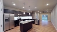 This single-story space showcases a thoughtfully designed open-concept layout that centers around a striking modern kitchen. Dark espresso cabinetry lines the walls from floor to ceiling, providing ample storage while creating a bold visual contrast against the white tile backsplash and light hardwood flooring that runs throughout. The kitchen is anchored by a central island topped with a dramatic veined marble-style countertop, offering additional workspace and a natural gathering point. A secondary countertop peninsula with a sink adds further functionality, complemented by a full suite of stainless steel appliances including a range, microwave, refrigerator, and dishwasher. Overhead, pendant lights hang above the island, while recessed lighting illuminates the space evenly. The open floor plan transitions naturally into an adjacent living area, where a ceiling fan and large glass door invite natural light and provide access to an outdoor area. The overall ambiance is clean, contemporary, and functional, with a cohesive design palette that balances dark and light tones effectively.