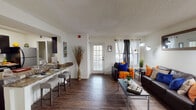 This single-floor residence offers a well-organized layout with dark hardwood-style flooring flowing throughout the main living areas, creating a cohesive and polished aesthetic. A welcoming entry hallway connects the various spaces, leading into an open living area bathed in natural light from multiple windows dressed with grommet-style curtains. The living room provides generous space for seating and is anchored by a textured ceiling and clean white walls.

The galley-style kitchen is a functional highlight, featuring dark espresso cabinetry, granite-style countertops, a double-basin sink, and black stainless appliances including a range and refrigerator. A breakfast bar with seating opens toward the living area, enhancing the sense of connectivity. A closet pantry adds practical storage.

Two bedrooms are each carpeted for comfort, with textured ceilings and ample wall space. Both bathrooms mirror the kitchen's design with matching granite-style countertops, vessel sinks, and tub-shower combinations with curtains, continuing the consistent material palette throughout.

A dedicated in-unit laundry closet with a top-load washer and overhead shelving adds everyday convenience. Overall, the residence presents a functional, cohesive design with consistent finishes throughout.
