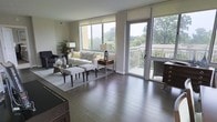 This well-appointed single-floor residence offers a seamless blend of comfort and modern design. The living area is anchored by rich, dark hardwood flooring that extends throughout, adding warmth and a sense of sophistication to the space. Large windows along the exterior wall flood the room with natural light, framing lush, tree-lined views that bring the outdoors in. A sliding glass door opens directly onto a private balcony, offering a serene outdoor retreat with open-air views of the surrounding greenery — an ideal spot for relaxing or entertaining. The living room is generously proportioned, providing ample space for comfortable seating arrangements and stylish furnishings. Neutral-toned walls complement the natural light and create a versatile backdrop for a variety of interior styles. The overall ambiance of the space is calm and inviting, with clean architectural lines and thoughtful finishes throughout. The combination of indoor openness and outdoor accessibility makes this residence a particularly appealing option for those seeking a bright, connected, and refined living environment.