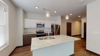 This single-floor unit presents a cohesive, modern design with wood-look flooring and neutral tones throughout. The kitchen serves as a central highlight, offering an open-concept layout anchored by a large island with a granite countertop and an undermount sink. Pendant lights with decorative shades hang above the island, adding a refined touch. Cabinetry features a two-tone design, combining white upper cabinets with dark lower cabinets, and granite countertops extend across the perimeter workspace. Stainless steel appliances, including a refrigerator, range, and over-the-range microwave, complete the kitchen's contemporary aesthetic.

The spacious living area benefits from natural light provided by large windows with vertical blinds, and the open floor plan creates a seamless flow between the kitchen and living spaces. An in-unit washer and dryer are also accommodated within the layout, adding convenience.

The bathrooms maintain the space's consistent design language. The full bathroom features a tub-shower combination, a dark-framed vanity mirror, and a single-sink vanity with a white countertop, while a separate half-bath provides additional functionality. Light neutral walls and tile flooring keep both bathrooms clean and understated.