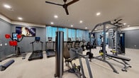 This fitness center in Vallejo, California offers a spacious, single-floor layout designed to accommodate a full range of workout needs. The space features a neutral color palette of gray and beige, with smooth carpeted flooring throughout and recessed lighting complemented by ceiling fans that ensure comfortable air circulation during workouts.

The cardio zone is well-appointed with multiple True-brand treadmills, stationary bikes, and additional aerobic machines, each equipped with individual monitors for entertainment or performance tracking. Large windows along one wall allow natural light to fill the space while providing exterior views, enhancing the overall workout environment.

The strength training area is equally comprehensive, featuring a multi-station cable and weight machine system, a dedicated free weights section with a full rack of dumbbells, and a bench press station. Floor-to-ceiling mirrors line the walls, amplifying the sense of space and allowing users to monitor their form during exercise.

A separate, clearly marked accessible restroom area is situated near the entrance, ensuring the facility is inclusive and convenient for all users. The overall design is clean, modern, and functional, with ample open floor space between equipment to allow for safe and comfortable movement throughout the gym.