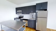 Located at 6300 City Avenue in Philadelphia, PA, this single-floor residence offers a clean, modern aesthetic throughout. The kitchen is a standout feature, showcasing contemporary dark cabinetry that extends floor to ceiling, providing ample storage space. A full suite of stainless steel appliances includes an electric range with oven, over-the-range microwave, dishwasher, and a full-size refrigerator. Light-toned countertops contrast elegantly against the dark cabinet finishes, while a classic white subway tile backsplash adds texture and visual interest to the space. A stainless steel prep surface provides additional workspace, making this kitchen as functional as it is stylish. Warm hardwood flooring runs throughout, adding a natural element that softens the modern, industrial feel of the kitchen. The open layout allows for easy movement and a sense of spaciousness. Natural light filters into the space, enhancing the bright, airy atmosphere created by the white walls and light surfaces. The overall design reflects a thoughtful balance of form and function, making this a highly practical and visually appealing living space.
