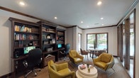 This single-level business center is thoughtfully designed to accommodate both focused work and casual collaboration. A striking floor-to-ceiling built-in bookcase anchors one wall, finished in rich dark wood and housing an extensive book collection alongside integrated computer workstations equipped with monitors and ergonomic task chairs. Crown molding and dark wood trim throughout the space lend a refined, professional character to the interiors.

The lounge area is furnished with a cluster of mustard-yellow leather accent chairs arranged around a tiered circular white coffee table, creating an inviting setting for informal meetings or quiet reading. A round dining-height table with upholstered chairs occupies a sun-drenched corner near the windows, ideal for small group gatherings or individual work sessions.

Expansive dark-framed windows and glass sliding doors line the perimeter of the space, flooding the interior with natural light and offering clear views of an outdoor courtyard and pool area beyond. The flooring transitions between hardwood-style planks and textured carpet, adding warmth and definition to different zones within the room. Recessed lighting throughout ensures consistent, comfortable illumination. The business center connects seamlessly to adjacent community spaces, making it a functional and welcoming hub for productivity.