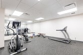 Located at 2 Quebec St in Guelph, Ontario, this fitness room offers a clean, functional space dedicated to health and wellness. The room features a drop-panel ceiling with recessed flat lighting that ensures bright, even illumination throughout. The flooring consists of durable speckled rubber gym matting, providing both safety and comfort underfoot during workouts.

The space is equipped with a selection of cardio and strength training equipment, including a treadmill, elliptical trainer, and stationary cycle. A weight rack holding a set of dumbbells, along with an adjustable workout bench, rounds out the strength training options. A full-length mirror along one wall visually expands the room and allows users to monitor their form during exercise.

A wall-mounted flat-screen television paired with a wall clock offers both entertainment and time management for users during their sessions. Motivational fitness-themed wall decals — featuring words such as "Fitness," "Endurance," "Wellness," and "Lifestyle" in a bold typographic design — add character and energy to the otherwise neutral, white-walled interior.

The room maintains a tidy and organized layout, maximizing the available floor space. A small waste bin and sanitizer dispenser near the entrance reflect attention to cleanliness and hygiene. Overall, this fitness room provides a practical and well-appointed environment for a complete workout routine.