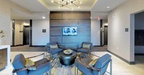 This single-floor lobby presents a well-appointed, contemporary design that balances functionality with sophisticated aesthetics. The main lounge area serves as the focal point, centered around a bold, wood-paneled accent wall housing a flat-screen television. A grouping of angular navy-blue chairs with patterned accent pillows surrounds a round, drum-style coffee table, all anchored by a vibrant geometric area rug in teal, gold, and grey tones. Overhead, a striking multi-arm chandelier with exposed bulbs adds a modern industrial touch to the tray ceiling above.

Adjacent to the lounge, a sleek reception-style console with dark wood paneling provides a practical yet elegant surface, flanked by framed abstract artwork. The foyer transitions smoothly into an entry corridor featuring large floor-to-ceiling windows and glass doors that flood the space with natural light and offer views to the exterior. Neutral, large-format tile flooring runs throughout, complementing the warm, taupe wall tones.

A dedicated mailroom alcove with built-in mailboxes is discretely positioned near the entrance, adding a practical element without disrupting the overall refined ambiance. The space as a whole conveys a welcoming, upscale character.