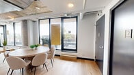 Located at 7 Fairbank Avenue West in Toronto's vibrant Corso Italia neighbourhood, this single-floor communal lounge offers a well-appointed, open-concept layout designed for gathering and relaxation. The space is defined by its industrial-modern aesthetic, most notably showcased through the exposed concrete ceiling with visible ductwork and HVAC systems running overhead — a striking architectural feature that adds an urban, loft-like character throughout.

Light oak hardwood flooring runs continuously across the entire floor, lending warmth and cohesion to the open plan. The living area is furnished with rattan-framed sofas and lounge chairs arranged around a central coffee table, creating an inviting seating zone. Floor-to-ceiling windows flood the space with natural light and frame panoramic views of the Toronto skyline and surrounding neighbourhood rooftops.

A sleek kitchenette lines one wall, outfitted with grey cabinetry, stainless steel appliances including a French-door refrigerator and dishwasher, and a clean white countertop with a sink — ideal for casual entertaining. A round dining table with chairs provides a dedicated dining area nearby.

Direct access to an outdoor terrace is available through a set of black-framed glass doors, extending the usable space outdoors and offering additional seating under the open sky.