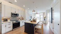 This single-floor residence is designed with a clean, contemporary aesthetic, blending functionality with modern style throughout its open layout. The kitchen serves as a central hub, featuring white flat-front cabinetry that extends to the ceiling, a textured tile backsplash in neutral tones, and a full suite of stainless steel appliances including a side-by-side refrigerator, range, and built-in microwave. A bold dark-finished island with a light countertop provides seating for multiple guests and acts as a striking contrast to the bright, white surroundings. Pendant lights with muted tones hang above the island, adding warmth to the space. The kitchen flows seamlessly into the living area, where large windows and a glass door flood the room with natural light and offer open views of the surrounding cityscape. Wide-plank light wood flooring runs throughout, unifying the space with a warm, cohesive tone. The bathroom continues the modern design language with large-format white wall tiles, a frameless glass shower enclosure with black hardware accents, and built-in storage nearby. Black hardware details appear consistently across the residence, lending a refined, intentional finish. The overall ambiance is bright, airy, and thoughtfully curated for contemporary living.