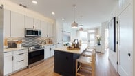 This single-floor residence is designed with a clean, contemporary aesthetic, blending functionality with modern style throughout its open layout. The kitchen serves as a central hub, featuring white flat-front cabinetry that extends to the ceiling, a textured tile backsplash in neutral tones, and a full suite of stainless steel appliances including a side-by-side refrigerator, range, and built-in microwave. A bold dark-finished island with a light countertop provides seating for multiple guests and acts as a striking contrast to the bright, white surroundings. Pendant lights with muted tones hang above the island, adding warmth to the space. The kitchen flows seamlessly into the living area, where large windows and a glass door flood the room with natural light and offer open views of the surrounding cityscape. Wide-plank light wood flooring runs throughout, unifying the space with a warm, cohesive tone. The bathroom continues the modern design language with large-format white wall tiles, a frameless glass shower enclosure with black hardware accents, and built-in storage nearby. Black hardware details appear consistently across the residence, lending a refined, intentional finish. The overall ambiance is bright, airy, and thoughtfully curated for contemporary living.