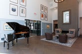 Located at 11115 Medlock Bridge Rd in Johns Creek, Georgia, this single-story space presents an elegantly designed interior that balances architectural grandeur with refined, curated details. The most commanding feature is the dramatic double-height ceiling in the main gathering area, which amplifies the sense of openness and draws the eye upward toward expansive floor-to-ceiling steel-framed windows. These large-pane windows flood the space with abundant natural light while framing picturesque views of the surrounding wooded landscape.

The flooring throughout is laid in a classic herringbone pattern, adding visual texture and a timeless aesthetic to the open layout. Crisp white brick walls contrast beautifully with soft blue-grey wainscoting panels, lending the space a polished yet welcoming character. A sleek linear fireplace set against a textured accent wall serves as a focal point in the lounge area, complemented by curated seating arrangements and layered area rugs.

A grand piano anchors one corner of the main hall, reinforcing the refined ambiance of the space. Framed artwork adorns the upper walls, adding warmth and personality. Steel-framed glass doors offer views into adjacent areas, including a bar and dining space with exposed beamed ceilings, enhancing the overall sense of connected, well-appointed interiors.