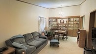 This single-story home presents an open-concept living and dining area that feels spacious thanks to its notably tall ceilings accented by crown molding. The living area is anchored by plush, comfortable seating arranged over neutral wall-to-wall carpeting, creating a relaxed and inviting atmosphere. A curtained window allows natural light to filter into the space, brightening the warm, cream-colored walls.

Transitioning seamlessly into the dining area, a classic chandelier hangs overhead, providing warm ambient lighting above a wooden dining table and chairs. The standout feature of this combined space is a large, built-in wooden shelving unit that spans an entire wall, offering generous display and storage space. The shelving showcases decorative items, books, and collectibles, adding a personalized and lived-in character to the room.

The textured ceiling adds a traditional architectural detail consistent throughout the space. Overall, the home conveys a cozy, functional aesthetic with classic design elements, making it a comfortable and well-utilized single-floor living environment.