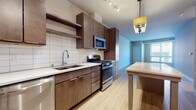 This single-floor space showcases a contemporary design with a cohesive blend of warm and neutral tones throughout. The kitchen serves as a central highlight, featuring rich dark-wood cabinetry extending floor to ceiling, a white subway tile backsplash, and light quartz countertops. A full suite of stainless steel appliances includes a gas range, built-in microwave, dishwasher, and refrigerator. A kitchen island with open shelving below provides additional prep space and storage, illuminated by a stylish pendant light fixture. Track lighting overhead ensures the workspace is well-lit.

Light hardwood-style flooring runs throughout the main living areas, lending warmth and continuity to the open-concept layout. The living area is generously proportioned, featuring clean white walls and a large window that draws in natural light, creating an airy and welcoming atmosphere.

The bedroom is finished with neutral carpet, soft walls, and a sizable window fitted with blinds, offering a comfortable and quiet retreat. The bathroom is finished with light tile flooring and includes a full soaking tub with shower, maintaining the unit's clean and functional aesthetic throughout.