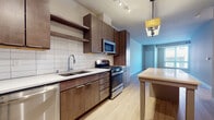 This single-floor space showcases a contemporary design with a cohesive blend of warm and neutral tones throughout. The kitchen serves as a central highlight, featuring rich dark-wood cabinetry extending floor to ceiling, a white subway tile backsplash, and light quartz countertops. A full suite of stainless steel appliances includes a gas range, built-in microwave, dishwasher, and refrigerator. A kitchen island with open shelving below provides additional prep space and storage, illuminated by a stylish pendant light fixture. Track lighting overhead ensures the workspace is well-lit.

Light hardwood-style flooring runs throughout the main living areas, lending warmth and continuity to the open-concept layout. The living area is generously proportioned, featuring clean white walls and a large window that draws in natural light, creating an airy and welcoming atmosphere.

The bedroom is finished with neutral carpet, soft walls, and a sizable window fitted with blinds, offering a comfortable and quiet retreat. The bathroom is finished with light tile flooring and includes a full soaking tub with shower, maintaining the unit's clean and functional aesthetic throughout.