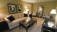 This single-floor space showcases a thoughtfully designed living area centered around a neutral-toned sectional sofa paired with coordinating black accent pillows. The room features warm olive-green walls that create an inviting, cozy atmosphere, complemented by plush wall-to-wall carpeting in a neutral beige tone. A sleek, dark-finished coffee table with glass inlay panels serves as the focal point of the seating area, accompanied by a patterned accent chair in black and white geometric print. An arched, dark-framed floor mirror adds visual depth and elegance to the space, while a glass-and-metal display cabinet showcases decorative accents. Soft ambient lighting from table lamps enhances the warm ambiance of the room. Two framed black-and-white artworks are displayed on the wall, adding a sophisticated artistic touch. A partial view into an adjacent room reveals a bright, open area with white cabinetry, suggesting additional functional living space. The overall design balances classic and contemporary elements through a consistent dark and neutral color palette, creating a refined, welcoming environment.