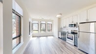 This 2-bedroom, 1-bathroom unit is thoughtfully designed to maximize functionality within a compact footprint. The open-concept living and kitchen area serves as the heart of the unit, where large black-framed windows spanning multiple walls flood the space with natural light and provide views of the surrounding neighborhood. Light wood-style plank flooring extends throughout the main living area, contributing to a clean, contemporary aesthetic.

The kitchen is efficiently outfitted with a full suite of stainless steel appliances, including a refrigerator, electric range, built-in microwave, and dishwasher. Two-toned cabinetry pairs white upper cabinets with gray lower cabinets, complemented by granite-style countertops, creating a polished, modern look.

Both bedrooms feature plush carpeting and receive ample natural light through large windows. The primary bedroom is a comfortable size and includes closet space. A walk-in closet with built-in shelving and a large window is also present, offering practical storage.

The bathroom features large-format tile flooring and a tiled walk-in shower with mosaic accent tile detailing, lending a clean and updated feel to the space.