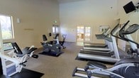 This fitness center offers a well-appointed workout environment designed to support a range of cardiovascular exercise routines. The space features soaring ceilings that lend an open, airy feel to the room, preventing any sense of confinement during workouts. The flooring is finished with durable, easy-to-maintain carpeting accented by protective rubber mats beneath the equipment.

The gym is outfitted with a solid selection of cardio machines, including treadmills, a recumbent stationary bike, an upright exercise bike, and additional aerobic equipment — all from reputable fitness brands. The machines are thoughtfully arranged to allow comfortable spacing between users.

Natural light filters in through a set of glass-paned double doors that connect to an adjacent lounge or common area, providing a welcoming view and easy access between spaces. A second set of exterior double doors offers direct entry and exit from the fitness room, adding convenience.

The walls are painted in neutral, light tones that contribute to the clean and motivating atmosphere of the space. An informational bulletin board is mounted near the entrance, and safety signage is clearly visible throughout. Overall, this fitness center combines functionality with a pleasant, open ambiance suitable for daily exercise.