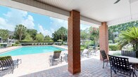 This outdoor pool area offers a well-appointed recreational space designed for both relaxation and leisure. A covered pavilion anchored by classic brick columns provides a shaded retreat, with a decorative brick-paver floor underfoot that adds a refined, structured aesthetic. The pavilion opens fully to the pool deck, creating a seamless transition between shaded and open-air areas.

The generously sized swimming pool features clear, inviting blue water and is surrounded by an expansive sundeck furnished with multiple lounge chairs and seating arrangements, accommodating both sunbathers and social gatherings. Umbrella-shaded seating along the far end of the deck offers additional relief from the sun.

The space is framed by mature, lush landscaping, including tall trees, manicured hedges, and flowering shrubs that lend a sense of privacy and natural beauty. The blooming crape myrtle trees add a vibrant pop of color to the greenery. The combination of the brick architectural elements, open sightlines, and verdant surroundings creates a serene, resort-inspired ambiance well-suited for outdoor enjoyment throughout the warmer months.