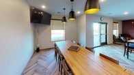 This well-appointed single-floor space seamlessly blends comfort and modern design across an open-plan layout. The living area serves as a cozy gathering spot, anchored by a built-in entertainment center with rich wood open shelving on either side, a wall-mounted flat-screen TV, and a mosaic tile fireplace surround that adds a decorative focal point. A plush sectional sofa, a live-edge wood coffee table, and a patterned area rug complete the welcoming atmosphere, while decorative woven wall art adds texture and character.

The fully equipped kitchen is a standout feature, showcasing warm walnut-toned cabinetry, granite countertops, and a glass mosaic tile backsplash. Stainless steel appliances — including a range, microwave, dishwasher, and refrigerator — offer full functionality, while a deep double basin sink and open wine rack add practical elegance. The eye-catching herringbone hardwood flooring ties the kitchen and dining areas together beautifully.

The dining area accommodates a generous long harvest-style wooden table beneath a trio of bold matte black and gold pendant lights, offering ample seating for group gatherings. A wall-mounted TV and access to a side door add convenience. Throughout the space, recessed lighting, neutral walls, and cohesive natural wood tones create a warm, inviting, and functional environment.