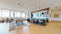 This contemporary social lounge in Folsom, CA is a well-appointed amenity space designed for gathering and entertainment. The interior is anchored by a striking bar area featuring a navy blue stone-topped island with dramatic veining, framed by warm wood accents and illuminated by a series of globe pendant lights in brass fixtures. White textured tile backsplash and open shelving complete the refined wet bar aesthetic, complemented by a wall-mounted TV and bar seating for multiple guests. Adjacent to the bar, a full-size pool table rests on a layered area rug, offering a central recreational focal point. The lounge seating area features sculptural chairs in mustard velvet, a boucle sofa, and a rich caramel loveseat arranged around a colorful area rug, all set against a striking slatted wood accent wall with an integrated large-format display. Floor-to-ceiling glass doors flood the interior with natural light while connecting seamlessly to an expansive outdoor terrace. The terrace is finished with large-format stone tile and offers multiple seating vignettes, including slatted wood sofas, a covered dining area with a built-in grill, and hanging egg chairs along the perimeter railing, all framing sweeping open-sky views.