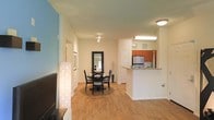 This single-story space is thoughtfully designed with an open-concept layout that seamlessly connects the living, dining, and kitchen areas. Warm hardwood flooring flows throughout, creating a cohesive and inviting atmosphere. The living area, positioned toward the front, provides a comfortable space for relaxation, while a striking teal accent wall adds a pop of color and visual interest.

The dining area sits centrally within the open floor plan, accommodating a round table and chairs beneath track lighting that illuminates the space effectively. A large framed mirror enhances the sense of depth and light in this area. Adjacent to the dining space, a full-length framed mirror further amplifies the bright, airy feel.

The kitchen is defined by warm wood cabinetry, a stainless steel refrigerator, and a peninsula with a breakfast bar that offers both additional counter space and casual seating. A large overhead fluorescent light ensures the kitchen is well-lit. The kitchen's open pass-through design maintains the connected, flowing nature of the overall layout. A hallway leads to additional rooms, completing this well-organized and functional single-floor space.