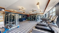 Located at 1100 Cricket Ln in East Walpole, MA, this well-appointed fitness center offers a spacious, single-floor layout designed to support a full range of workout routines. The interior is styled with bold navy blue accent walls, warm orange trim details, and a crisp white ceiling, creating a modern and energizing atmosphere. Plaid-patterned carpet tile flooring runs throughout, adding texture while providing a comfortable surface underfoot.

The cardio zone is positioned along the large windows that line one wall, allowing natural light to pour in and offering pleasant outdoor views of the surrounding greenery — an uplifting backdrop for treadmill and rowing machine workouts. Multiple ceiling fans are strategically placed overhead to maintain airflow throughout the space.

The strength training area is equally well-equipped, featuring a multi-functional cable machine, a full dumbbell rack with a wide range of weights, an adjustable workout bench, and a collection of stability and medicine balls. A wall-mounted flat-screen display adds a multimedia element to the space.

Decorative wall art, including a large map mural and framed prints, contributes to the room's polished aesthetic. Recessed and pendant lighting, combined with the abundant natural light from the oversized windows, ensures the space is well-lit at all hours. Glass-paneled entry doors provide easy access and visual connectivity to the exterior.