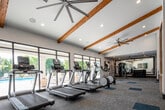 This fitness center is designed to support a comprehensive workout experience across multiple distinct zones. The main gym floor is expansive and airy, featuring soaring ceilings accented by exposed wooden beams and large ceiling fans. Patterned carpet tiles in neutral and teal tones cover the floor, complementing the bright, welcoming atmosphere. This area is outfitted with treadmills, ellipticals, stationary bikes, and a variety of strength machines. Floor-to-ceiling windows and glass doors line the far wall, offering direct views of an outdoor pool and patio, flooding the space with natural light.

A dedicated functional training studio is enclosed by sleek black-framed glass partition walls and features warm wood-plank flooring, a ribbed ceiling with recessed lighting, and ample mirror coverage. This studio connects seamlessly to the main floor while maintaining its own distinct environment.

A separate, more intimate training room is outfitted for high-intensity and boxing-style workouts, featuring a multi-functional wall-mounted rack loaded with resistance bands, TRX straps, kettlebells, medicine balls, a punching bag, and a mini trampoline. This room is finished with dark teal walls and wood flooring, creating a focused, energizing atmosphere.

The entry area features vaulted ceilings with beam detailing, a bold adventure-themed graphic display, and package lockers visible through glass-panel walls.