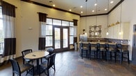 This single-floor communal space blends functionality with a warm, inviting aesthetic. The open layout is anchored by rich slate tile flooring in earthy tones, which grounds the room and adds a sense of permanence and texture underfoot. Tall ceilings with crown molding and recessed lighting lend an airy, spacious quality to the interior, while dark wood trim throughout provides a refined contrast to the cream-toned walls.

A standout feature is the set of French doors dressed with two-tone brown and white curtains, flooding the room with natural light and offering direct access to an outdoor balcony with views of surrounding greenery. This seamless indoor-outdoor connection enhances the overall ambiance of the space.

The kitchenette area is well-appointed with a stainless steel refrigerator, a decorative tile backsplash, and a dark countertop bar with cushioned bar-height seating — ideal for casual gatherings. Pendant lights hang above the bar, adding warmth and a focal design element. Decorative wall art in a grid arrangement adds a curated touch to the kitchen wall.

A casual dining area with a round table and dark chairs sits near the French doors, offering a comfortable spot for informal meetings or socializing. The overall atmosphere is polished yet approachable, making this a versatile and well-designed communal gathering space.